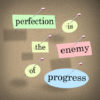 Perfection is the Enemy of Progress quote or saying with words on pieces of paper pinned to a bulletin board to illustrate you should not wait to take action until things are perfect